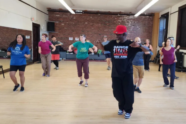 Man in black clothing and red hat dances in a dance studio with several students behind him